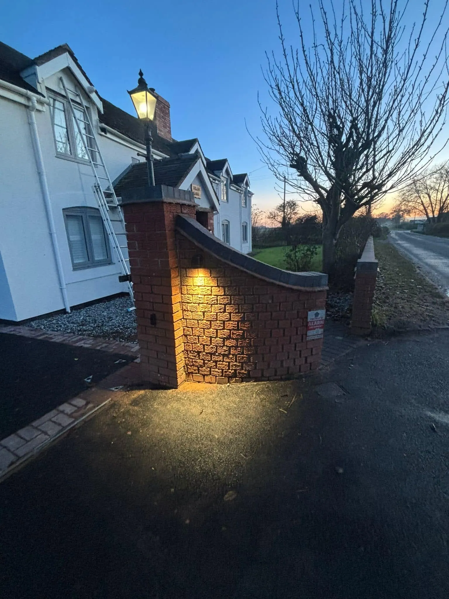A driveway entrance at dusk lit by a lamp post and a wall-mounted uplighter